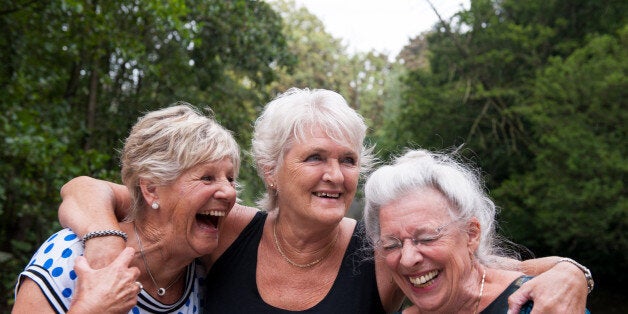 Three elderly women holding one another and laughing out loud, outdoors in a green environment on an overcast day.