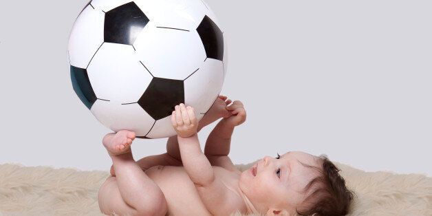 Little baby boy, lying naked on her back on woolen fleece; holding big soccer ball, on white background
