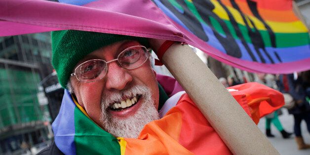 Artist Gilbert Baker, designer of the Rainbow Flag, is draped with the flag while holding a banner that reads