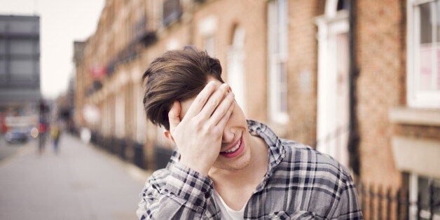 close portrait of a young stylish man covering his eyes with his hand, while smiling joyfully, in front of a wide perspective of a long street disappearing in a graduate blur