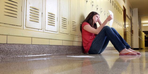 A middle school girl sitting in hallway of school looks upset while looking at her classwork.