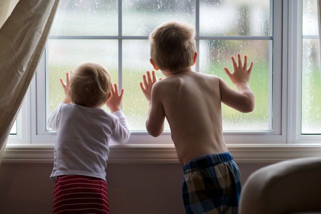 Two young children press their faces up against window looking outside on rainy day.