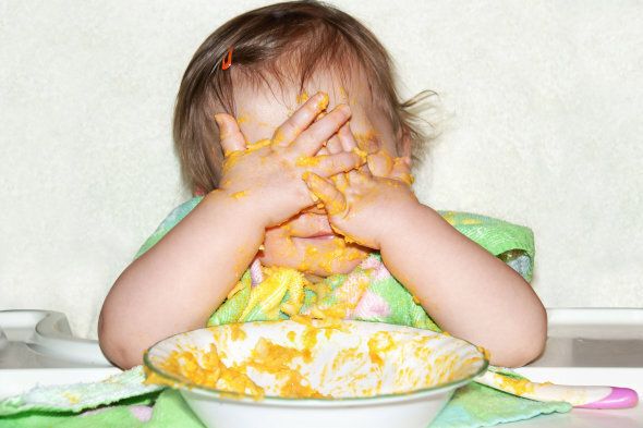 Funny baby with food covered face by putting her hands over her eyes to play picaboo during mealtime, wrong time to do so.