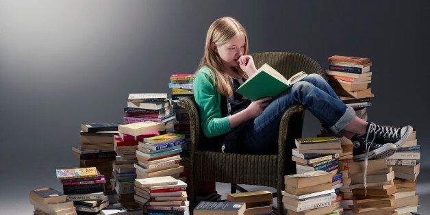 teenage girl sat in chair reading book surrounded by piles of books