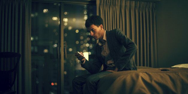 Serious young businessman sitting on a bed of a hotel room and using his smart phone. San Francisco lights in a background
