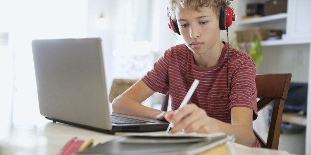 Young boy making notes on writing pad while using laptop and headphones at table