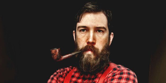 A man dressed as a lumberjack with a plaid shirt and suspenders has a big beard and a pipe in his mouth.