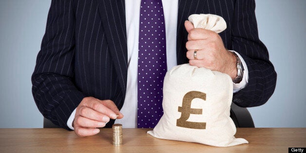 A British banker sat at a desk holding a money bag and adding a pound coin to a stack of coins.