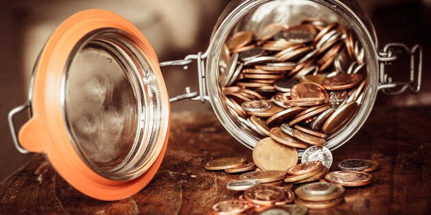 A jar of sterling coins is tipped on its side with the coins spilling out on to a wooden surface. The jar denotes a savings pot.