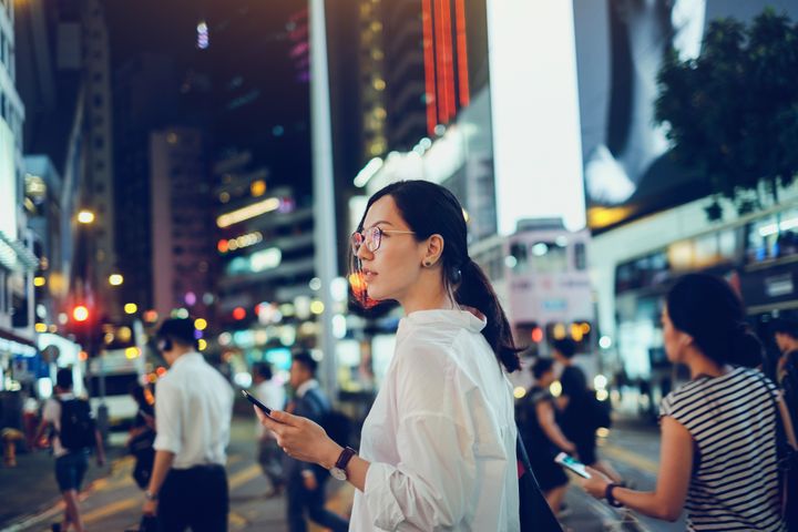 Beautiful Asian woman using mobile phone while crossing road in busy downtown city street at night