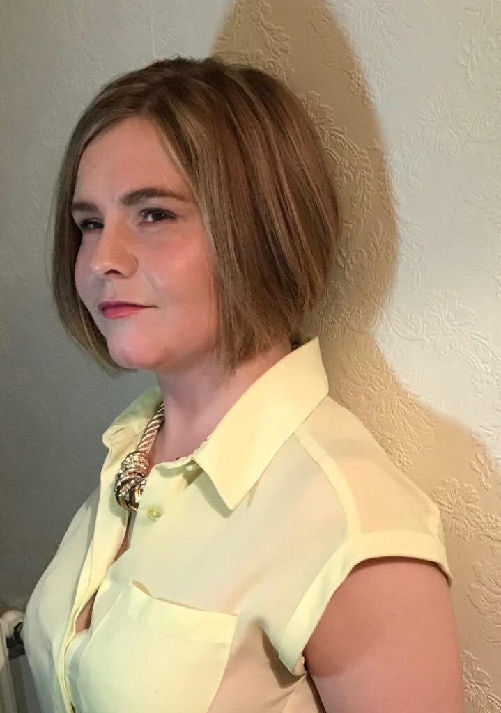 A side-shot of Sassy Wyatt in a yellow blouse wearing a gold necklace. She has short blond hair and is looking directly at the camera and smiling.