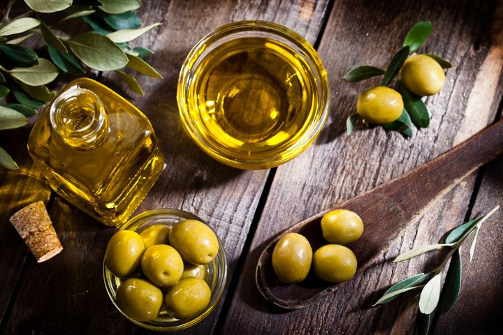 Top view of an olive oil bottle and a little glass bowl filled with green olives on rustic wood table. Two olives with leaves are at the top-right while a bowl filled with olive oil is at the center-top beside the two olives. An olive tree branch is at the left-top corner. A wooden spoon with three olives comes from the right. Predominant colors are gold, green and brown. DSRL studio photo taken with Canon EOS 5D Mk II and Canon EF 100mm f/2.8L Macro IS USM