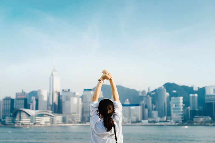 Rear view of woman traveller enjoying her time in Hong Kong, taking a deep breath with hands raised against Victoria Harbour and city skyline