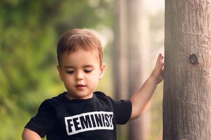 Brunette toddler plays in a tree lined park while wearing a feminist shirt.