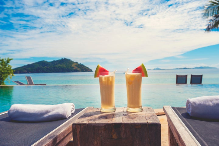 Cocktails by the pool with beach in the background. There are sun lounges with towels. The drinks are Pina coladas with fruit. Copy space