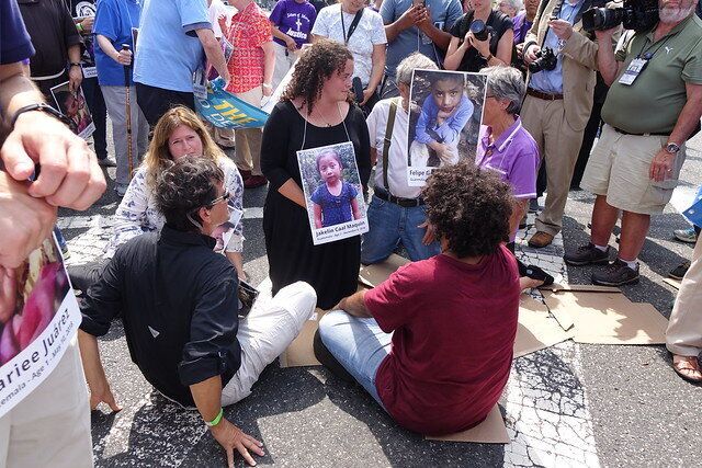 Jacqueline Small, a postulant with the Benedictine Sisters of Erie, kneels on a crosswalk during a Catholic protest in Newark while wearing a photo of Jakelin Caal Maquin, a migrant child who died in detention.