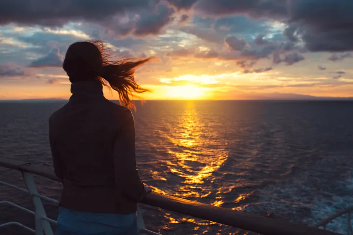 Rear view of a young woman standing by the fence of a sailing ship and enjoying the sunset view.