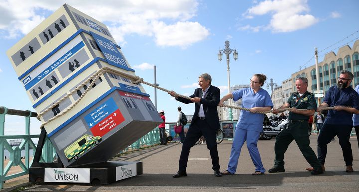 Dave Prentis with members of the NHS as they pretend to pull back a mock up of a hospital falling over on the sea front in Brighton