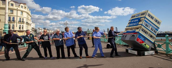 NHS workers pretend to pull back a mock up of a hospital falling over on the sea front in Brighton