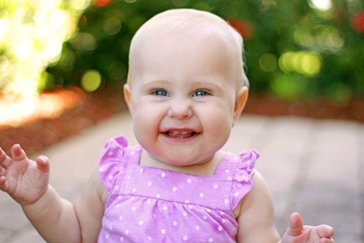 A super happy 10 month old baby girl with new front teeth is smiling as she sits in the garden on a summer day.