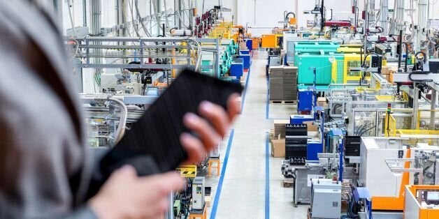 Horizontal color image of businesswoman - unrecognizable person - working with digital tablet in large futuristic factory. Woman standing on top of a balcony, holding touchpad and checking inventory of a manufacturing company on touchscreen tablet. Focus on futuristic machines in background, businesswoman's hands holding black tablet defocused.