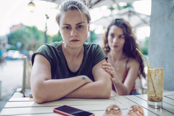 Two young women sitting sad and irritated on a coffee table; one of them is trying to console the other.