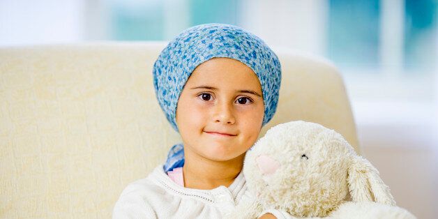 A hopeful little girl with cancer, sitting at home with her stuffed animal, she is smiling and looking at the camera.