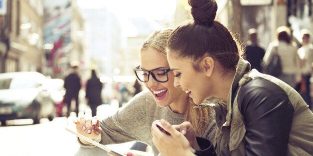 Two young women using a digital tablet. They are sitting on a city street. One woman has dark hair that is pulled into a bun on top of her head. She has on a tan jacket with black sleeves. She is wearing blue jeans and has a gold-colored watch on her left wrist. The other woman has blond hair that is parted in the middle. She is wearing black glasses, a gray shirt and blue jeans. The blond woman is holding the tablet in her left hand. Both women are smiling. Cars and people are blurred in the background.