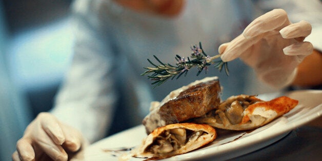 Closeup tilt of blurry female chef placing rosemary on a steak meal before serving. She's using protective gloves when dealing with ready to eat food.