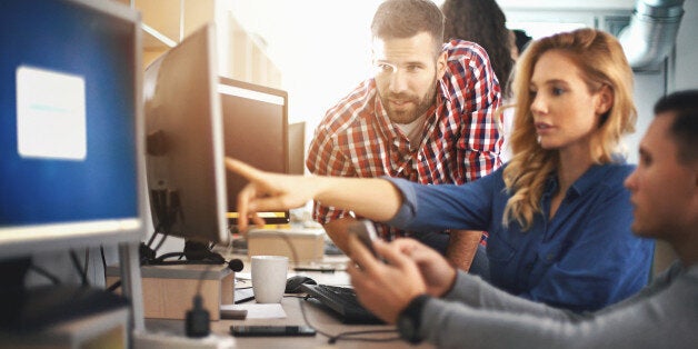 Closeup of group of application developers discussing about certain code for an application. There are two men and a woman and she's pointing at computer screen. Blurry people in background, also released. Orange light flaring from the background.