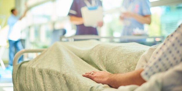 a female senior nurse or matron is chatting to a young a young student nurse is attending to the dressings trolley at the foot of a patient's bed . They are all defocussed apart from the patient's hand in the foreground.