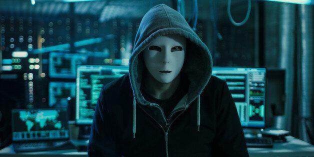Dangerous Internationally Wanted Hacker with Covered Face Looking into the Camera. In the Background His Operating Room with Multiple Displays and Cables.