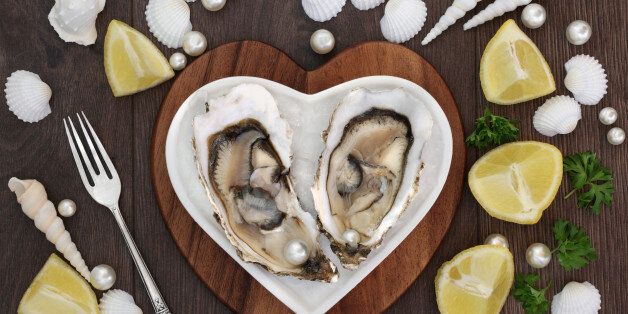 Oysters and pearls on a heart shaped plate on a maple wood board with lemon fruit, parsley, shells and an old silver fork over old oak background.