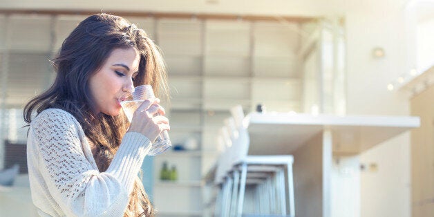 Woman drinking a glass of water. She is sitting on the terrace outside her luxury home. Kitchen in the background. She is smiling. Copy space right hand side
