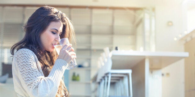 Woman drinking a glass of water. She is sitting on the terrace outside her luxury home. Kitchen in the background. She is smiling. Copy space right hand side