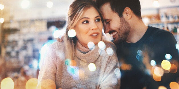 Closeup of happy mid 20's couple having fun at local coffee place on winter night, somewhere around Christmas time.They are sitting by a window side by side and laughing. The woman is looking through the window. Both wearing warm casual clothing. Tilt shot,toned image.