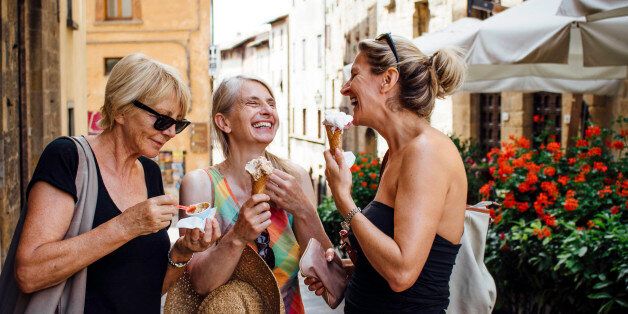 Three mature female friends standing eating Italian ice-creams while in a street in Tuscany during summer. They are smiling and facing each other and enjoying their holiday.