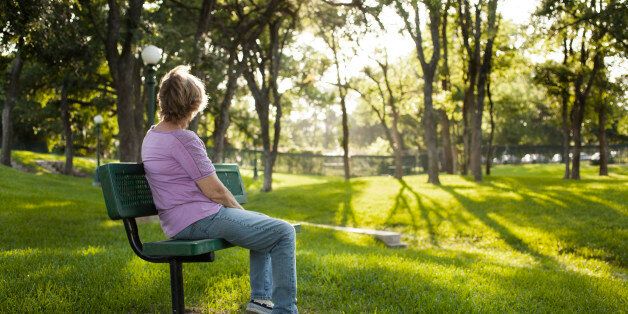 Rear view of one mature woman sitting on a park bench in the summer season. Bright sunny day with lush green grass and trees. The woman gazes off into the distance as she relaxes on a beautiful day. Solitude, lonliness, contemplation. She has short blond hair and wears a purple shirt and jeans. Copyspace to right in this tranquil nature scene.