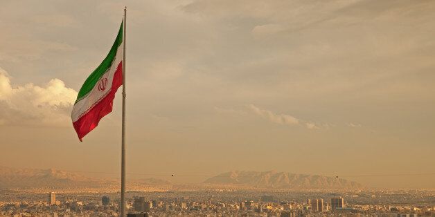 Iran flag waving in the wind above skyline of Tehran lit by orange glow of sunset.