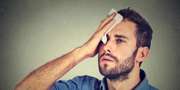 Tired man stressed sweating having fever headache isolated on gray wall background. Worried guy wipes sweat on his face