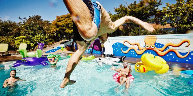 Man in mid air jumping into outdoor pool during party with friends swimming and cheering in background