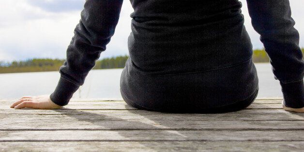 Rear view closeup photo of woman in black clothes sitting on a wooden pier looking out over the lake.