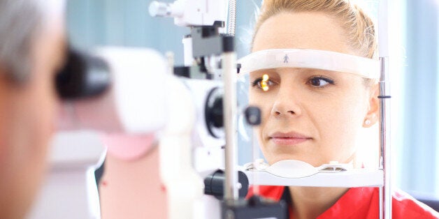 Closeup of late 20's blond woman visiting optician. She placed her head into phoropter machine while senior male doctor is examining her eyesight. The woman has brown eyes and wearing red t-shirt.