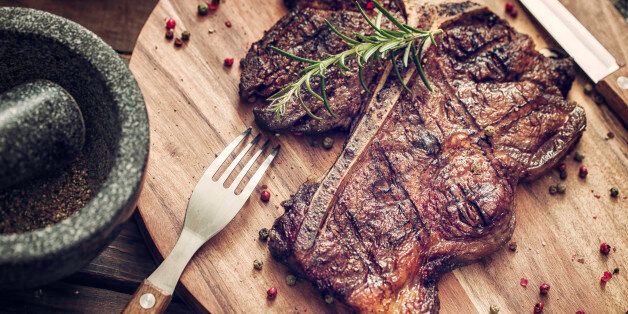 Delicious T-Bone Steak Medium Roasted with Herbs and Pepper. The steak is on a wooden plate served with knife and fork.