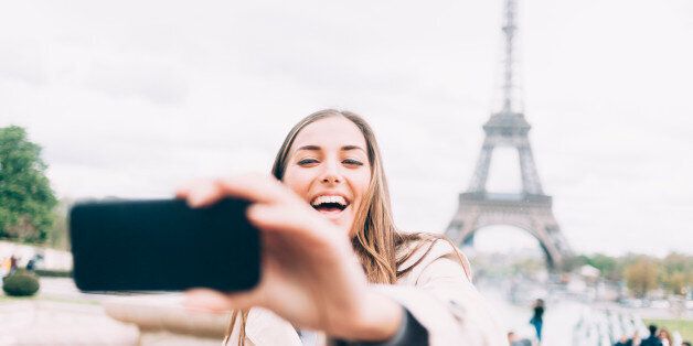 Laughing young woman making selfie in front of the Eiffel tower. With long hair and wears coat. Street view, tourists and a park on background.