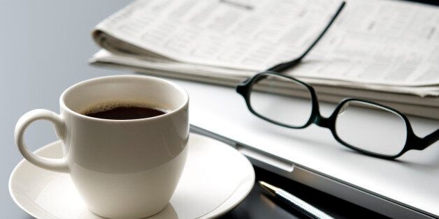 A close up of a desk at a business office. This desk includes a cup of black coffee served in a white cup on a white saucer, a laptop computer, a pair of glasses and newspaper.