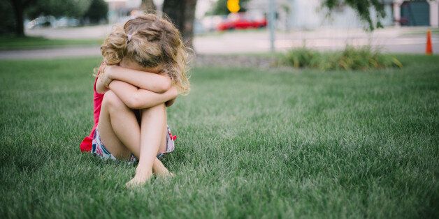 Little girl with curly hair sits on grass and cries into her arms.