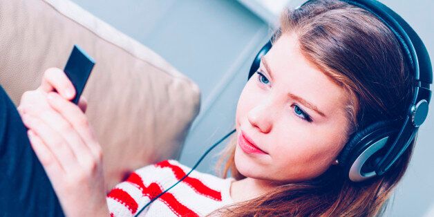 Teenage girl sits in a sofa with a device to play music and podcasts. The item is blurred and could pass both as a MP3 player and a smart phone. She wears headphones which are connected to the audio player. The girl is around 14 years old. She has brown hair and wears a blouse with red and white stripes.