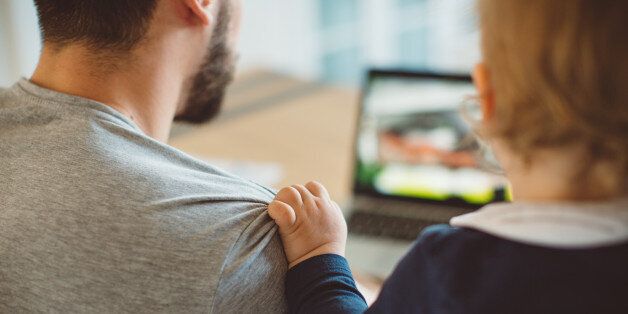 Young man sitting at the table in a kitchen, working on laptop, while baby holding him for shoulder. Paying bills online. View from behind.