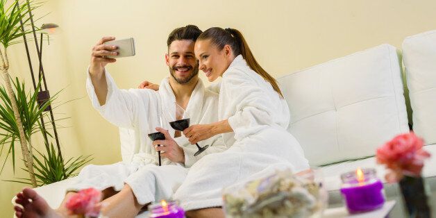 Couple in love enjoying wellness weekend and taking Selfie. They are sitting in a sofa , holding wine glasses, embracing and smiling.
